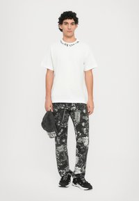 Camiseta blanca de manga corta con texto negro, pantalones negros con un patrón de diseños abstractos, y una gorra gris en la mano con detalles del logo.