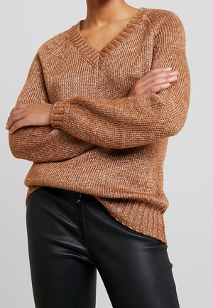 Person iført en brun strikket V-hals sweater med lange ærmer foldet over brystet, kombineret med sorte læderbukser.