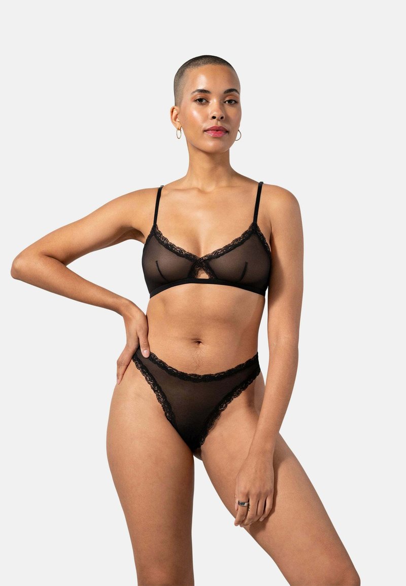 Zwarte kanten lingerieset met een doorzichtige bralette en bijpassende thong met delicate geschulpte randen. Model poseert tegen een effen achtergrond.