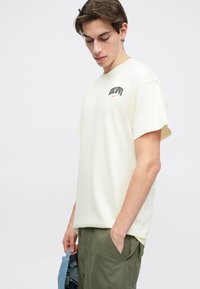 Krēmu krāsas kokvilnas T-krekls ar brīvu piegriezumu, kas rotāts ar melnu grafiku un orandžu logo uz krūšu. Apvienots ar haki biksēm.