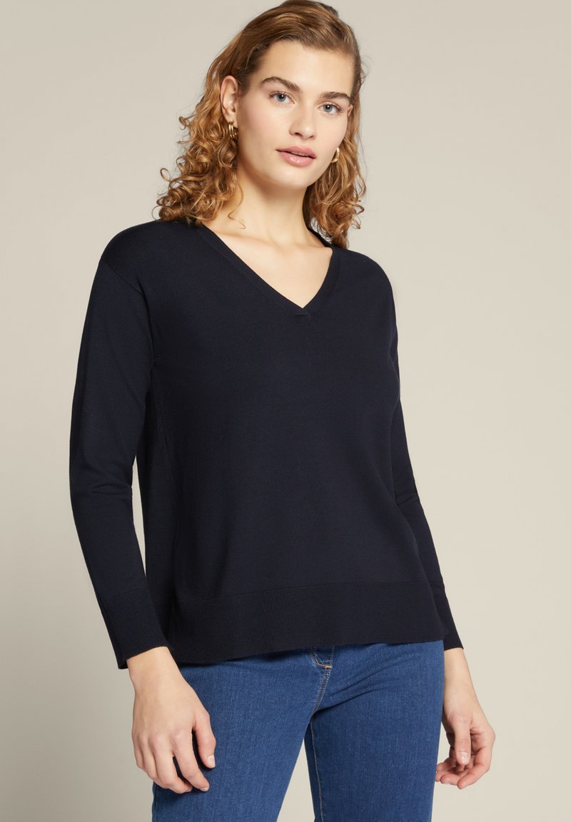 Donkerblauwe v-hals sweater met lange mouwen, gemaakt van zachte stof. Iets ontspannen pasvorm, geribde manchetten en zoom, gedragen met blauwe jeans.
