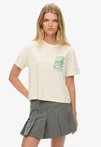 Cremefarvet kortærmet T-shirt med grøn grafisk tryk, der viser en bygning og tekst. Sammen med en grå stribet nederdel.