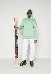 Μέντα πρασινή κουκούλα, λευκά παντελόνια χιονιού, μαύρες μπότες σκι, κρατώντας ροζ και σκούρα σκι με πρόσδεση. Περιλαμβάνει λογότυπο στην κουκούλα και την μπιτζάνα.