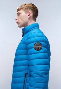 Giacca a vento blu con design a trapuntato, colletto alto e zip laterale. Presenta una patch rotonda con logo sulla manica sinistra. Texture liscia.