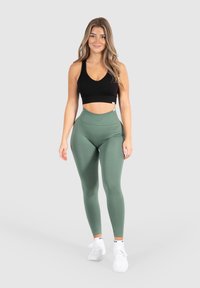Černý sportovní crop top, vysoké olivově zelené legíny, hladká textura, přiléhavý střih, doplněno bílými teniskami. Neutrální pozadí.