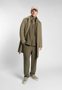Olijfgroene outfit met een lange jas, turtleneck, overhemd met knopen en bijpassende broek, aangevuld met een beige pet en lichte sneakers.