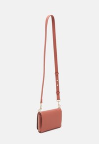 Borsa a tracolla piccola in pelle di corallo testurizzata con tracolla regolabile e zip e hardware in tonalità oro, appesa su uno sfondo bianco semplice.