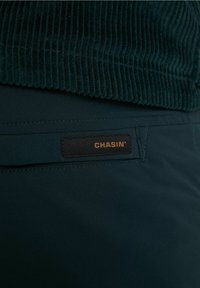 Mörkgrön kordtov med en överdel som går över i ett slätt mörkt tyg nedanför. Har en svart etikett med "CHASIN’" i guldtext.