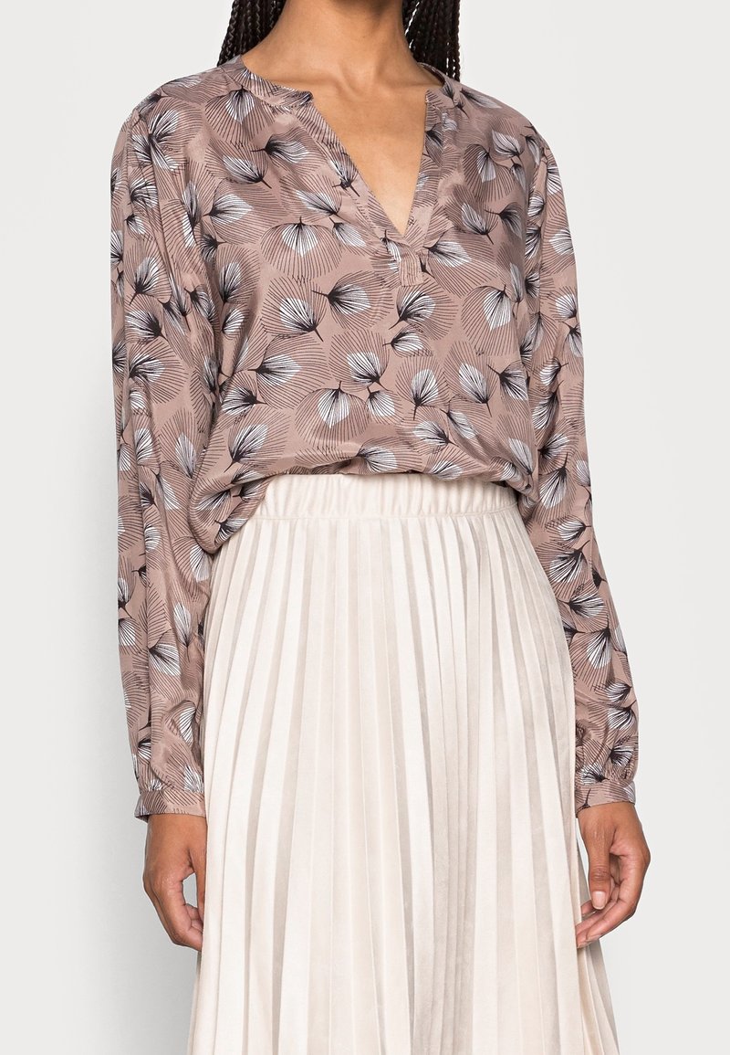Femme portant un chemisier mauve à fleurs à manches longues rentré dans une jupe plissée blanche taille haute sur un fond clair.