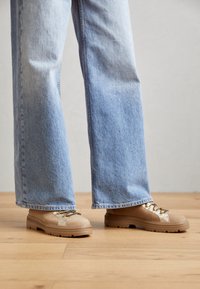 Beige afslappede sko med teksturerede overflader, metalliske snørebåndsdetaljer og solide gummisåler, parret med lyseblå brede bukser.