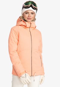 Pink skijakke med hætte, justerbar kant og lynlås-lommer. Hvide handsker med branding, båret over et lyse farvet outfit.