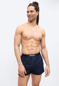 Homme musclé avec des dreadlocks portant un boxer bleu marine BOSS, debout et souriant en regardant de côté sur un fond uni.