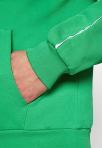 Sweatshirt verde com um tecido texturizado. Apresenta um bolso frontal e um detalhe branco ao longo da manga. Close-up de uma mão no bolso.