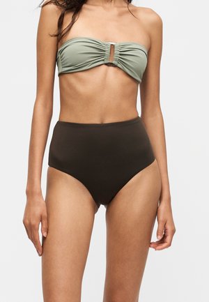 Femme portant un maillot de bain deux pièces avec un haut bandeau vert olive et un bas taille haute marron foncé sur un fond uni.