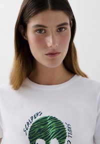 T-shirt bianca con un design grafico verde e blu di un teschio con un motivo a strisce zebrate. Collo rotondo e materiale in cotone morbido.
