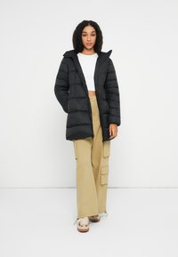 Černá péřová bunda s kapucí, s prošívaným designem, předními kapsami a zipem. Nositá přes bílý crop top a béžové cargo kalhoty.