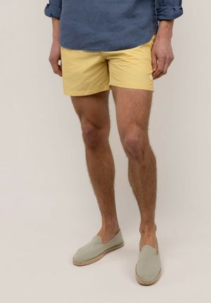 Hombre con pantalones cortos amarillos, camisa azul remangada y alpargatas beige sin cordones de pie contra un fondo liso.
