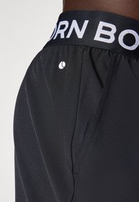 Schwarze Sportshorts mit einem strukturierten elastischen Bund, der mit "BORN" beschriftet ist. Verfügt über ein kleines Logo an der Seite und eine glatte Stoffoberfläche.