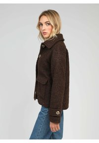 Femme aux cheveux blonds portant une veste texturée marron foncé avec de grands boutons et un jean bleu, de profil contre un fond uni.