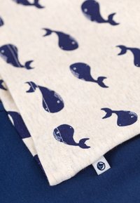 Λευκό ύφασμα με σχέδιο ναυτικού φά λαινα σε στρώσεις πάνω από σκούρο μπλε ύφασμα, με μια μικρή ετικέτα με λογότυπο στην άκρη.