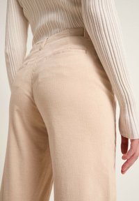 Beiga manchesterbyxor med ribbad struktur, med hög midja och bakfickor. Modellen bär en gräddfärgad ribbad tröja.