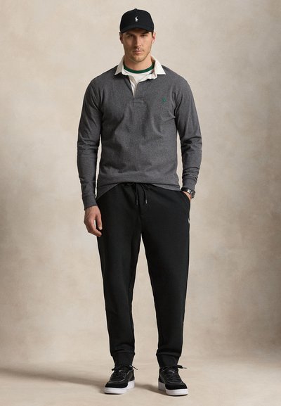 Mand iført en grå polotrøje med lange ærmer, sorte joggingbukser, sort kasket og sorte sneakers med hvide såler, stående mod en ensfarvet baggrund.