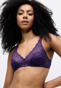 Bralette in pizzo di un profondo viola, con scollatura a onde e spalline, caratterizzato da motivi floreali intricati e da una texture morbida e trasparente.