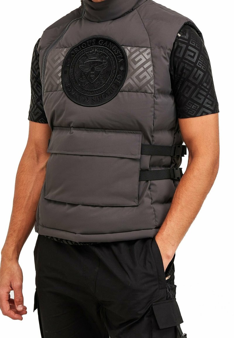 Gilet grigio imbottito con colletto alto, caratterizzato da un grande distintivo in rilievo, tasche frontali e cinghie nere. Indossato sopra una camicia nera a fantasia.