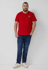 Rotes Baumwoll-T-Shirt mit kurzen Ärmeln und einem kleinen Design auf der Brust. Kombiniert mit blauen Jeans aus Denim und weißen Schnürschuhen.