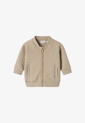 Giacca trapuntata beige con zip frontale, maniche raglan, polsini a costine e colletto rotondo. Presenta tasche laterali e un motivo a zigzag testurizzato.
