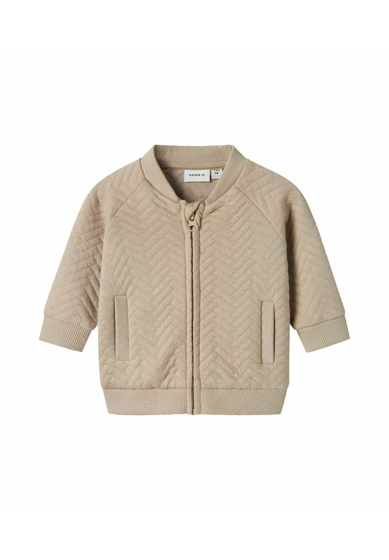 Giacca trapuntata beige con zip frontale, maniche raglan, polsini a costine e colletto rotondo. Presenta tasche laterali e un motivo a zigzag testurizzato.