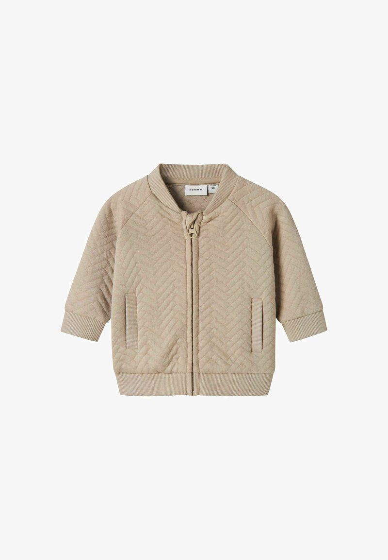 Beige gesteppte Jacke mit Reißverschluss vorne, Raglanärmeln, gerippten Bündchen und rundem Kragen. Mit Seitentaschen und einem strukturierten Chevron-Muster.