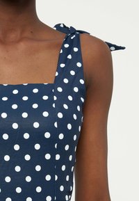 Κοντινό πλάνο ενός ατόμου με σκουρόχρωμο δέρμα που φοράει μπλε-ναυτικό φόρεμα με λευκές πουά και ιμάντες που δένονται στους ώμους, σε ουδέτερο φόντο.