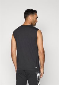 Férfi ujjtalan fekete sportpólóban és fekete nadrágban, fehér oldalsávokkal, háttal áll egy sima háttér előtt.