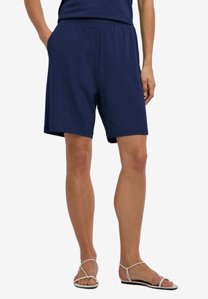 Osoba nosząca granatowe spodenki z elastycznym pasem i białe płaskie sandały na paski, stojąca z jedną ręką w kieszeni.