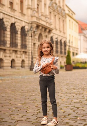 Child wearing a floral long-sleeve shirt, gray ribbed leggings, and multicolored sneakers, holding a tan leather crossbody bag. Cobblestone background.