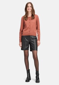 Kardigan w kolorze rdzy z okrągłymi guzikami, zestawiony z czarnymi szortami z ekoskóry. Modelka nosi czarne, kryjące rajstopy oraz botki na obcasie.
