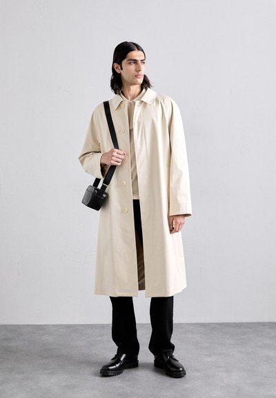 Bezs színű, klasszikus gallérral, elülső gombokkal és oldalsó zsebekkel készült pamut trench coat, fekete nadrággal és vastag talpú fekete cipővel párosítva.