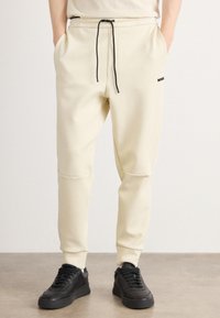 Cremfarbene Sweatpants aus weichem Stoff, mit einem Kordelzug in der Taille, schwarzen Akzenten und seitlichen Taschen, die an den Knöcheln schmal zulaufen.