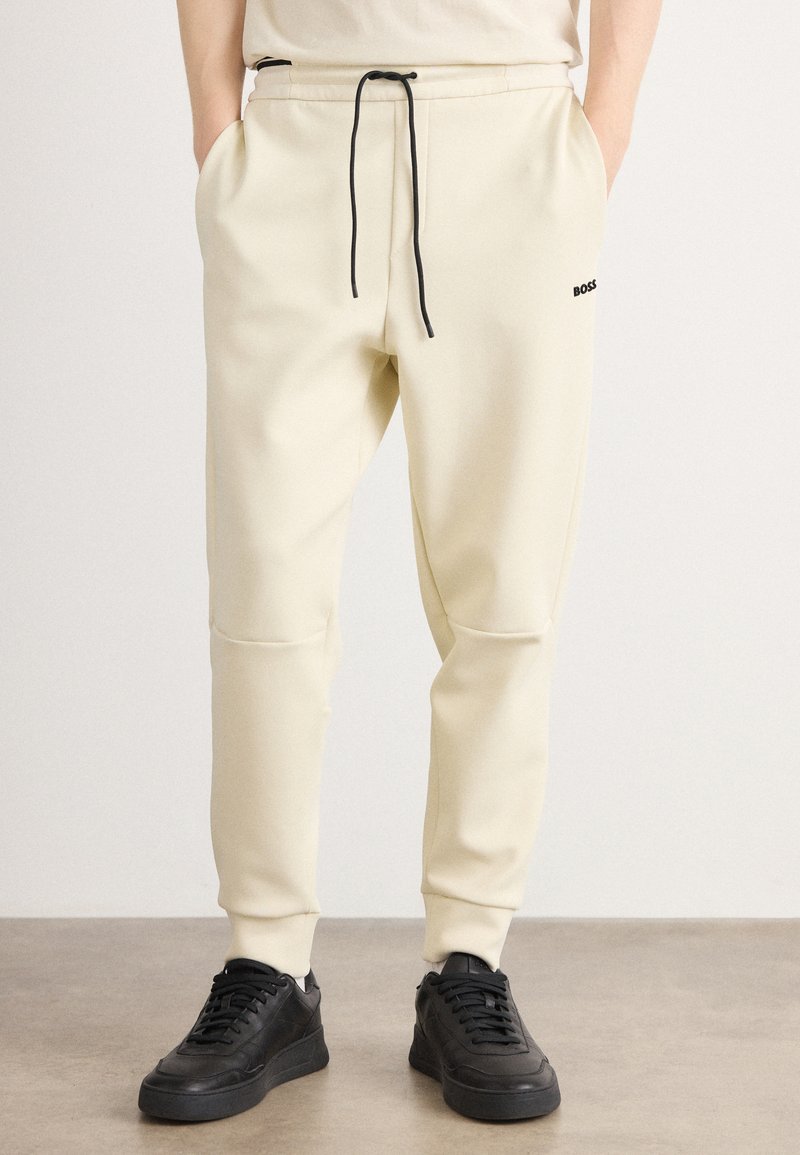 Cremfarbene Sweatpants aus weichem Stoff, mit einem Kordelzug in der Taille, schwarzen Akzenten und seitlichen Taschen, die an den Knöcheln schmal zulaufen.