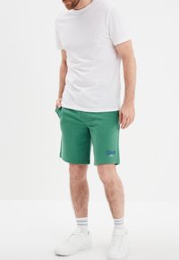 Bermudas de mezcla de algodón verdes con bolsillos laterales, cinturilla elástica y detalle del logo en morado. Combinadas con una camiseta blanca sencilla y zapatillas blancas.