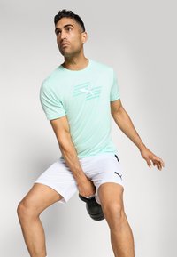 Lys mintgrøn atletisk t-shirt med korte ærmer, der har et hvidt stribe-logo. Båret med hvide shorts, mens der holdes en sort kettlebell.