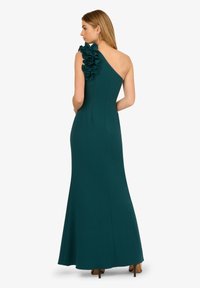 Jednoduché tyrkysové šaty na jedno rameno s volánkovým detailem, přiléhavým vrškem a rokajovou sukní, vyrobené z hladkého, elastického materiálu. Viditelné jsou průhledné podpatky.