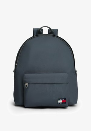 Mochila de tela gris con forma redondeada, correas de hombro acolchadas, bolsillo frontal con cremallera y un parche de logo visible en colores contrastantes.