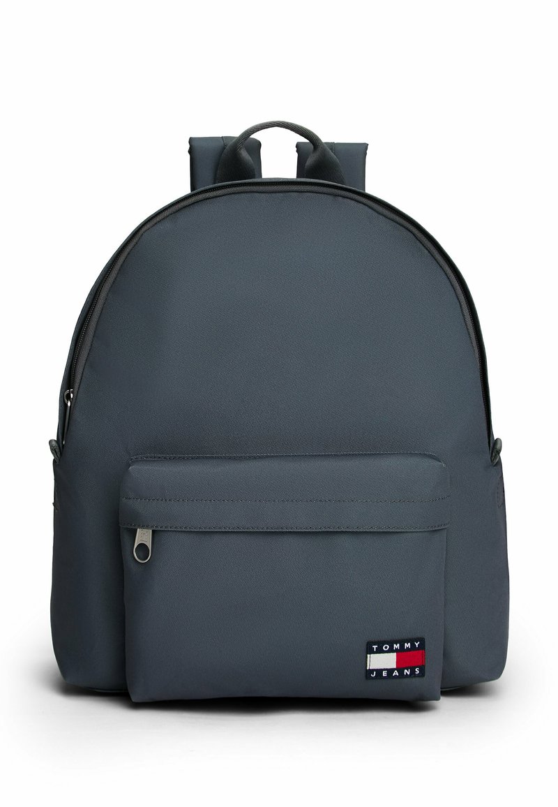 Gray fabric backpack with a rounded shape, padded shoulder straps, front zip pocket, and a visible logo patch in contrasting colors.