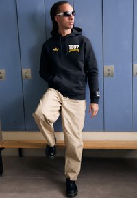 Svart hoodie med "1897 Juventus" i gult, beigefärgade byxor och svarta platåskor. Sportiga solglasögon kompletterar outfiten. Bakgrunden har skåp.