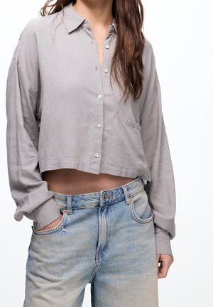 Kvinde iført en lysegrå, kort cropped skjorte med knapper og lyseblå, højtaljede jeans med den ene hånd i lommen.