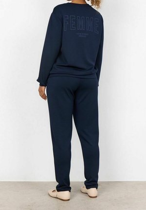 Femme portant un sweat-shirt et un pantalon bleu marine, avec le mot "FEMME" imprimé dans le dos du sweat-shirt, debout contre un mur blanc.