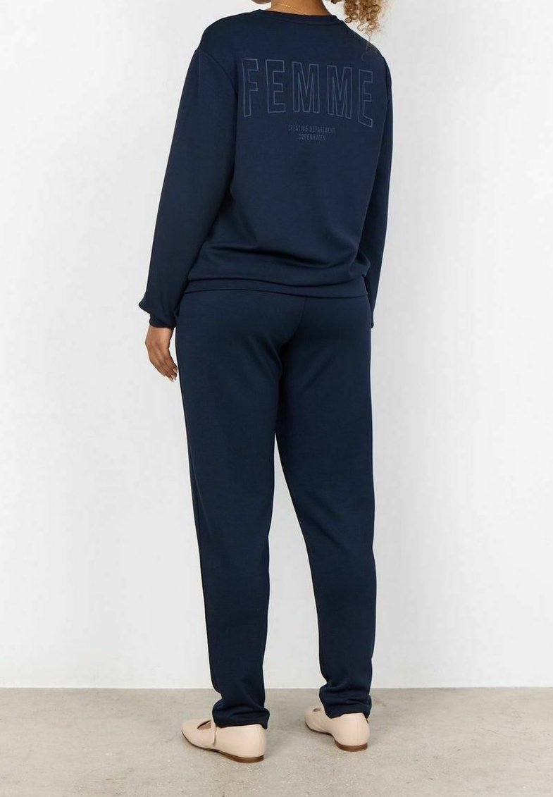 Femme portant un sweat-shirt et un pantalon bleu marine, avec le mot "FEMME" imprimé dans le dos du sweat-shirt, debout contre un mur blanc.