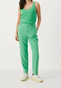 Groene mouwloze top en op maat gemaakte groene broek van gladde stof. Witte sneakers met een beige zool. Eenvoudig, modern ontwerp met een relaxte pasvorm.